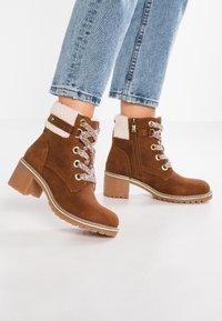 Bottines marron avec une finition en suède, doublure moelleuse, lacets décoratifs, œillets dorés et un talon épais. Fermeture éclair sur le côté pour un enfilage facile.