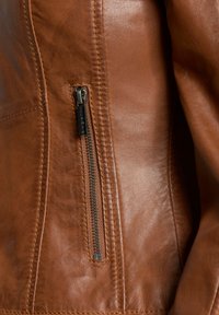 Braune Lederjacke mit glatter Textur, ausgestattet mit einem seitlichen Reißverschluss und sichtbaren Nähdetails. Der Reißverschluss hat eine metallische Oberfläche.