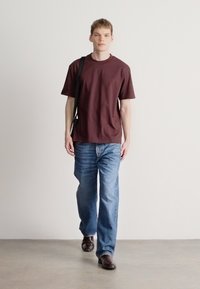 Krojena majica kratkih rokavov v maron rjavi barvi, modre kavbojke in temno rjavi čevlji. Majica ima sproščen kroj, enostavno obliko in je iz bombažnega materiala.