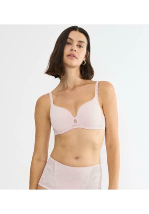 Vrouw met donker schouderlang haar, gekleed in een lichtroze gewatteerde beha en bijpassend kanten hoge tailleslipje, tegen een effen achtergrond.