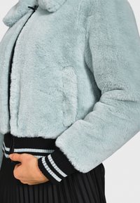 Giacca in pelliccia sintetica azzurro chiaro con texture soffice, dotata di cintura e polsini a coste neri con strisce bianche e tasche laterali.