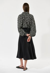 Črna cvetlična bluzo s pufastimi rokavi, kombinirana s črno satensko srednje dolgim krilom. Model nosi črne ravne sandale, pogled zadaj.