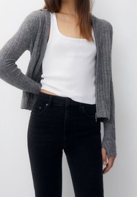 Cardigan gris de canalé con mangas largas, diseño abierto en la parte delantera, llevado sobre una camiseta blanca sin mangas y jeans negros de cintura alta. Textura suave.