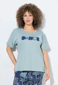 Svetle modré tričko s krátkým rukávem s texturou písmen "DANCE" v námořnické modři. V kombinaci s modrými kalhotami s potiskem v paisley vzoru.