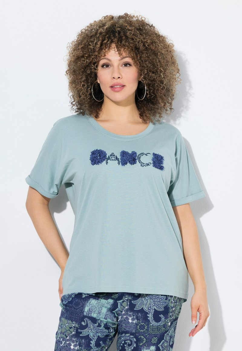 Svetle modré tričko s krátkým rukávem s texturou písmen "DANCE" v námořnické modři. V kombinaci s modrými kalhotami s potiskem v paisley vzoru.
