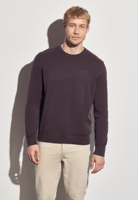 Dunkellila Rundhals-Pullover aus feinem Strickstoff, mit gerippten Bündchen und Saum. Kleines Logodetail auf der Brust. Kombiniert mit hellen Hosen.