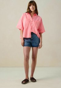 Pink button-up shirt with short sleeves and two front pockets, paired with blue denim shorts and brown sandals with wide straps.