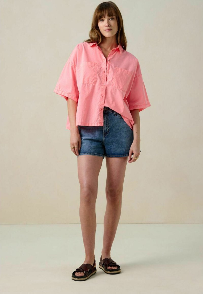 Pink button-up shirt with short sleeves and two front pockets, paired with blue denim shorts and brown sandals with wide straps.