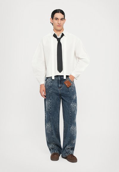 Camisa blanca recortada con lazos, corbata negra, jeans de mezclilla azul con bordado floral y zapatos marrones con cordones.