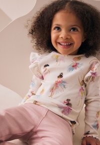 Wit geribd langarmshirt met kleurrijke prints van balletdanseressen; roze katoenen broek. Opvallend geruffleerde schouderdetail.