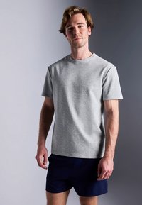 Harmaa lyhythihainen t-paita pyöreällä pääntiellä, pehmeästä kankaasta valmistettu, yhdistetty tummansinisiin shortseihin rentossa asennossa neutraalia taustaa vasten.