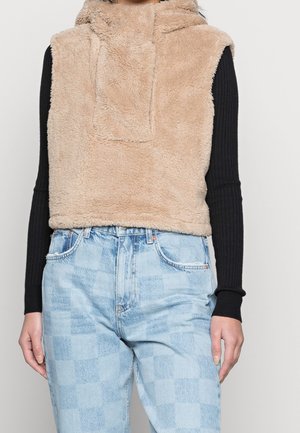 Femme portant un sweat à capuche beige duveteux sans manches par-dessus un haut noir à manches longues et un jean à carreaux bleu clair taille haute.