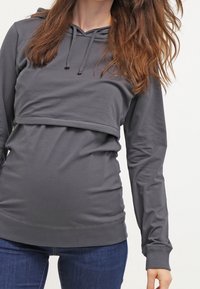 Felpa grigia a maniche lunghe con design corto, dotata di cappuccio con cordino e texture arricciata in vita, abbinata a jeans blu.