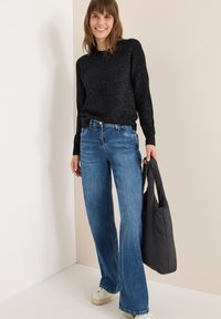 Czarny sweter dzianinowy, jasnoniebieskie szerokie dżinsy i kremowe sneakersy. Trzyma czarną pikowaną torbę. Prosty, casualowy strój na neutralnym tle.