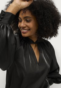 Blusa nera con scollatura a V e design plissettato. Presenta maniche a palloncino con dettagli a bottone. Texture liscia e leggermente lucida.