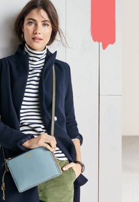 Manteau marine sur un col roulé rayé, tenant un petit sac à main bleu clair avec un logo doré, associé à un pantalon olive.