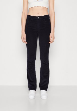Jeans évasés en velours côtelé marine avec fermeture à bouton et poches avant, associés à des baskets blanches, sur fond blanc uni.