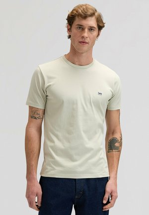 Giovane uomo che indossa una maglietta Lee color beige chiaro e jeans blu scuro, in piedi davanti a uno sfondo grigio uniforme con tatuaggi visibili sulle braccia.