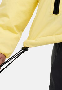 Giacca gialla con una texture liscia e leggera. Presenta cordini neri regolabili all'orlo e polsini elasticizzati per una vestibilità ottimale.