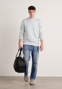 Sudadera de cuello redondo azul claro, jeans rectos desgastados, zapatillas grises y un bolso negro cruzado, caminando frente a un fondo neutro.
