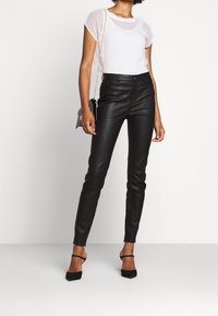 Černé kalhoty z umělé kůže s úzkým střihem a vysokým pasem, doplněné průhledným bílým topem s krátkými rukávy. Modelka má na nohou černé lodičky na podpatku.
