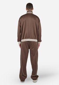 Survêtement marron avec une texture satinée. Veste zippée avec un col montant et des finitions en ivoire. Pantalons à coupe ample avec un passepoil ivoire.