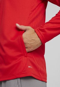 Rood atletisch shirt met lange mouwen en een textuurpatroon, met een verborgen zak aan de zijkant en een "DRYCELL" label nabij de zoom.