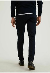 Marineblauwe slim-fit broek met een gladde textuur, voorzien van een discrette tailleband en achterzakken, gecombineerd met zwarte leren schoenen.