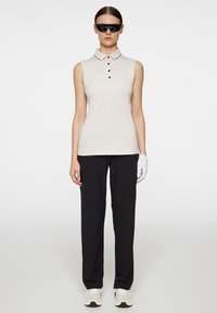 Ärmloös polo i ljus beige med krage och fem svarta knappar, i kombination med svarta byxor och en vit golfhandske.