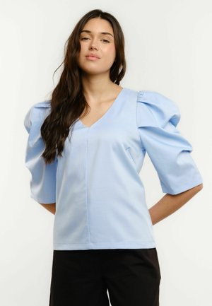 Young woman with long dark hair wearing a light blue puff-sleeve blouse and black pants, standing with hands behind her back.