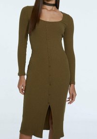 Robe côtelée vert olive avec manches longues, décolleté carré, boutons sur le devant et une fente en bas. Le matériau semble extensible et moulant.