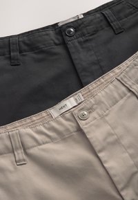 Dos pares de pantalones de algodón para hombre, uno negro y uno beige, doblados con botones visibles, trabillas para cinturón y una etiqueta de ropa en el par beige.