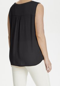 Haut noir sans manches avec un détail plissé dans le dos, tissu léger, ourlet arrondi et texture lisse, porté avec un pantalon de couleur claire.