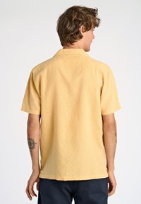 Uomo dai capelli castano chiaro che indossa una camicia gialla a maniche corte e pantaloni scuri, visto di schiena su uno sfondo bianco semplice.