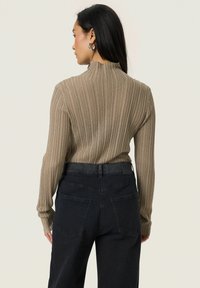 Kvinde med langt, mørkt hår iført sølvfarvede hoops, en beige ribbet sweater og sorte højtaljede jeans, vendt væk mod en ensfarvet baggrund.