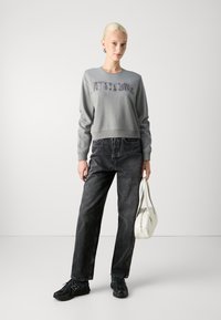 Hallakrüütatud hall sweatshirt tekstuuri kirjadega, mustad teksapüksid ja mustad tossud. Hoides valget käekotti, minimaalne disain.
