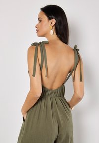Olive green romper with adjustable tie straps, open back, and smocked waist. Lightweight fabric features a textured finish.