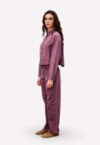 Femme debout dans une veste en denim violet et un pantalon taille haute assorti, portant des chaussures marron, faisant face à gauche sur un fond blanc.