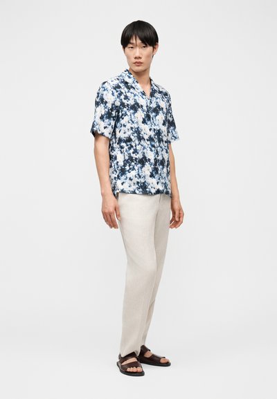 Άνδρας με μπλε και άσπρο λουλουδάτο κοντομάνικο πουκάμισο, μπεζ παντελόνι και σκούρα καφέ σανδάλια, στέκεται μπροστά σε απλό φόντο.