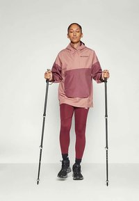 Let lette lyserøde og burgunderrøde jakke med en frontlomme, parret med burgunderrøde leggings. Personen holder vandrestave og har sorte sko på.