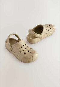 Beige Plastikclogs mit einem Riemen, ausgestattet mit Belüftungsöffnungen, einer strukturierten Sohle und einem runden Zehendesign. Leicht zu reinigen und leichtgewichtig.