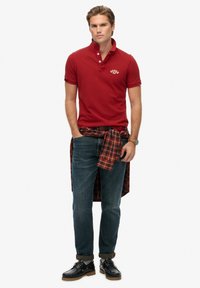 Polo rouge avec patte à trois boutons, jean bleu foncé, et chemise à carreaux marron nouée autour de la taille. Chaussures noires avec semelle marron clair.