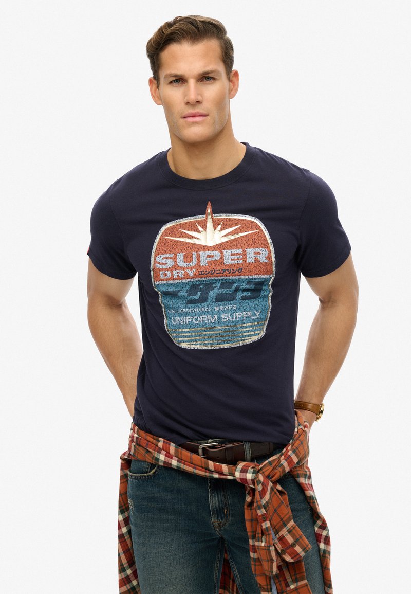 Camiseta de algodón azul marino con un gran estampado gráfico en naranja y azul, etiquetada "SUPER DRY UNIFORM SUPPLY", con cuello redondo y mangas cortas.