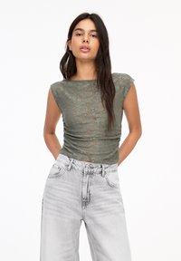 Olijfgroene kanten top met kapmouwen, gerimpelde details en doorschijnende textuur, gedragen met lichtgrijze denim jeans met hoge taille.