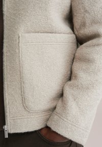 Jachetă din fleece texturat, de culoare alb-murdar, cu închidere cu fermoar și buzunar aplicat frontal. Manșetele mânecilor sunt finisate cu o cusătură curată.