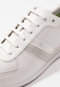 Bílé kožené tenisky s šedými semišovými akcenty, plochými šněrovadly a logem na boku. Mají vysoce pohodlnou vložku.