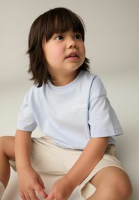 T-shirt di cotone azzurro chiaro con maniche corte, caratterizzata da un logo bianco sulla parte anteriore, abbinata a pantaloni corti beige. Tessuto morbido, vestibilità rilassata.