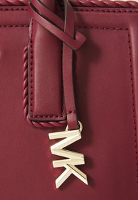 Burgundy läderhandväska med flätade accenter och en logocharm i guldton. Slät textur med sömnadsdetaljer längs kanterna.