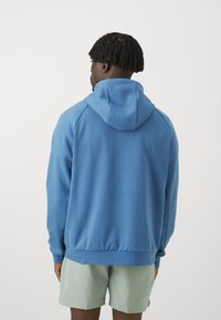 Blue hoodie with a spacious hood, ribbed cuffs and hem, made of soft material. Paired with light green shorts in a casual setting.