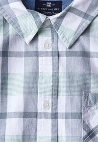 Košile s krátkým rukávem s kostkovaným vzorem v světle zelené a šedé barvě. Obsahuje zapínání na knoflíky, špičatý límec a náprsní kapsu. Vyrobena z měkké tkaniny.